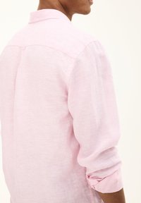 Camicia di lino rosa chiaro con vestibilità comoda, colletto a camicia, maniche arrotolate e una texture morbida. Presenta un yoke posterior per un tocco di dettaglio in più.
