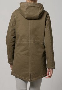 Olivgrön parkajakett med foder, huva, långa ärmar och kontrasterande vit sömnad. Har en längre rygg och snedslits.
