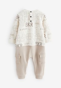 Ensemble bébé deux pièces beige comprenant un haut blanc avec des motifs de texte marron et un pantalon beige avec deux poches latérales et des poignets aux chevilles.