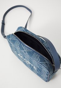Borsa a tracolla in denim con una tracolla in pelle blu, caratterizzata da un motivo geometrico, zip argentata e un logo bianco sul davanti.