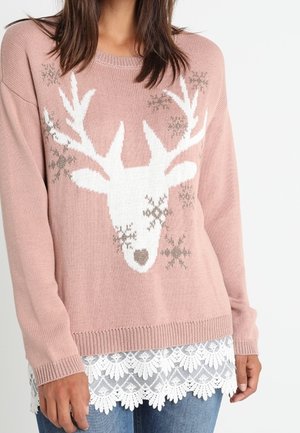 Žena v růžovém svetru s motivem bílého soba a vloček, doplněném modrými džíny a bílým krajkovým lemem.