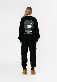 Sudadera negra con un gráfico de dibujos animados en la espalda, combinada con pantalones cargo negros y botas color marrón. La tela parece suave; el diseño es casual.