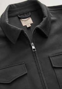 Chaqueta negra con cremallera hecha de tela texturizada, que presenta un cuello, dos bolsillos en el pecho y una cremallera metálica suave. La etiqueta dice "NEXT."