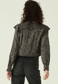 Blouse noire avec un motif de feuilles blanches, épaules volantes, tissu texturé, manches longues, portée avec un jean foncé, mettant en valeur une coupe décontractée.