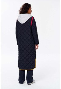 Černý prošívaný dlouhý kabát s šedou mikinou, červenými akcenty na rukávech a žlutým lemem na spodním okraji. Zadní pohled ukazuje uvolněný střih a designové detaily.