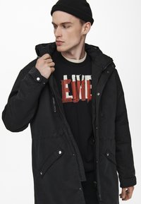 Svart parka-jacka med dragkedja och tryckknappar, med huva, justerbara ärmslut och framfickor. Bärs över en grafisk t-shirt.