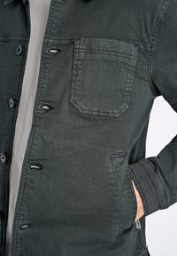 Chaqueta de mezclilla verde con cierre de botones en la parte delantera, un bolsillo en el pecho y bolsillos laterales. Cuenta con costuras sutiles y un cuello suave.