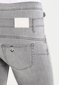 Jeans de denim gris con una superficie texturizada, una etiqueta de marca blanca en la cinturilla y detalles distintivos en los bolsillos con herrajes plateados.