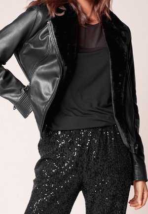 Femme portant une veste en cuir noire avec col en fourrure, un haut noir transparent et un pantalon noir à sequins, debout avec une main sur la hanche.