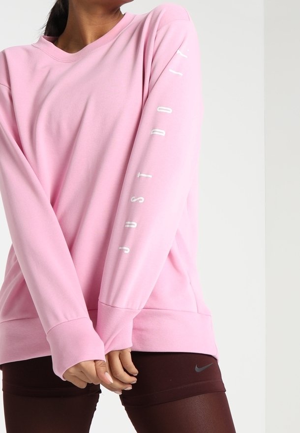 Sudadera rosa de manga larga en tejido suave con cuello redondo. Presenta "JUST DO IT" en blanco en la manga derecha. Combinada con pantalones cortos oscuros.