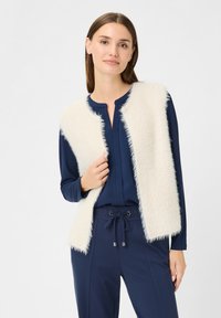 Faux-Fur-Weste in Creme mit einer lockeren Textur, über einem marineblauen Oberteil getragen. Sie hat einen runden Ausschnitt und einen entspannten Schnitt, ergänzt durch marineblaue Hose.