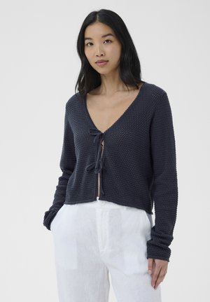 Kobieta w ciemnogranatowym dzianinowym kardiganie z wiązaniami z przodu i białych, wysokich spodni, stojąca na tle jednolitego jasnego tła.