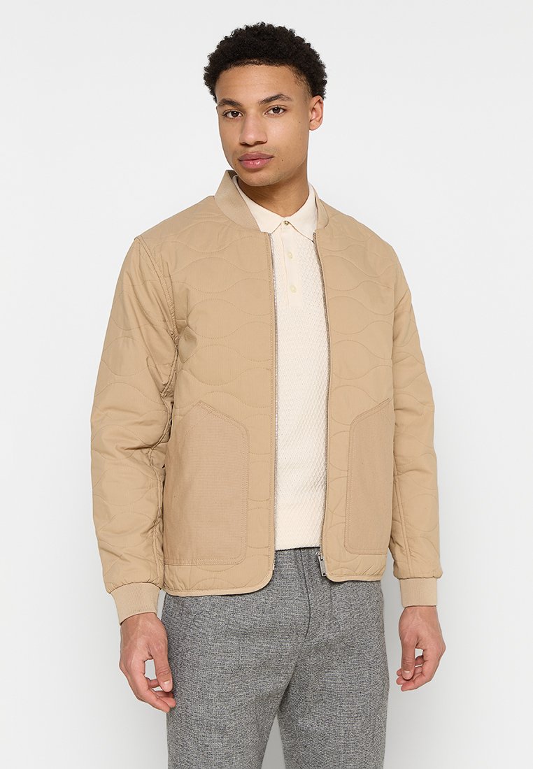 Giubbotto bomber beige trapuntato con colletto tondo, zip frontale e tasche applicate. Indossato sopra un polo chiaro e pantaloni grigi.