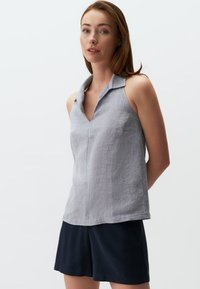 Top senza maniche in lino grigio chiaro con colletto e scollatura a V. Indossato con pantaloni corti navy scuri, il tessuto ha una finitura testurizzata.