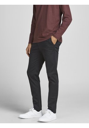 Persona con pantalones oscuros de corte ajustado, zapatillas blancas y una camisa de manga larga color granate, con una mano en el bolsillo.