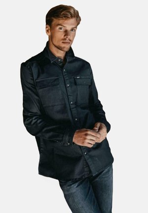 Donkerblauwe overhemdbloes met twee borstzakken, lange mouwen en een gladde textuur, gecombineerd met donkere denim jeans. Geen zichtbare patronen.