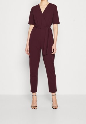 Vrouw draagt een bordeauxrode korte mouw overslagjumpsuit met een taillekoord en zwarte sandalen met hoge hak en enkelriem tegen een effen achtergrond.