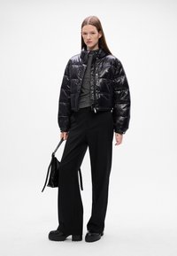 Giacca puffer nera, lucida e crop, con colletto alto, abbinata a pantaloni neri a gamba larga e calzature nere chunky. Tiene una piccola borsa nera.