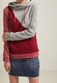 Felpa con cappuccio grigia e rossa, con un design diagonale e texture a contrasto. Presenta lacci e una tasca frontale; abbinata a una gonna di jeans scura.