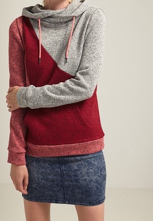 Felpa con cappuccio grigia e rossa, con un design diagonale e texture a contrasto. Presenta lacci e una tasca frontale; abbinata a una gonna di jeans scura.