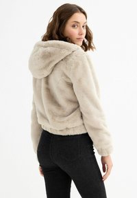 Chaqueta con capucha de imitación piel en beige claro con diseño corto. Presenta una textura suave y dobladillo acanalado. Combinada con vaqueros oscuros de talle alto.