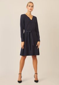 Abito lavorato a maglia navy con maniche lunghe, scollo a V, vita cinturata, gonna a ruota e texture liscia; abbinato a tacchi neri con cinturino alla caviglia.