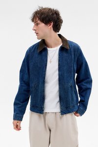 Ung mand iført en blå denimjakke over en hvid t-shirt og beige bukser, som kigger til venstre mod en ensfarvet baggrund.