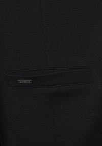 Černá tkanina se strukturovaným vzorem obsahuje diskrétní kapsu na zip a kovový štítek značky, zdůrazňující čisté linie a minimální design.