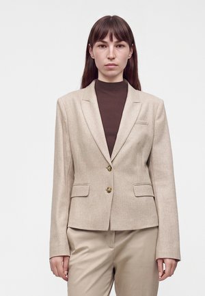 Femme aux cheveux bruns raides portant un blazer beige sur un col roulé côtelé marron foncé et un pantalon beige, debout devant un fond clair uni.