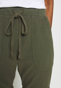 Pantalons vert olive avec une taille à cordon de serrage, dotés de poches latérales et d'un tissu doux et texturé. Un nœud visible accentue la taille.