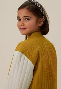 Gequilted jas met mosterdgele en crèmekleurige panelen, voorzien van een zonborduursel; model heeft een dierenprint haarband.