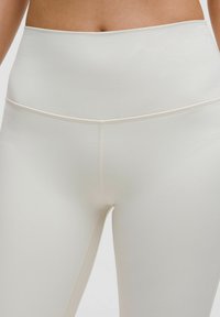 Legginsy z wysokim stanem wykonane z gładkiego, elastycznego materiału w kremowym kolorze. Charakteryzują się minimalistycznym wzornictwem i subtelnym przeszyciem wzdłuż szwów.