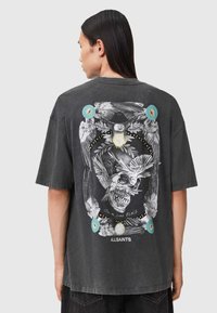 Melna izmēra t-kreklu ar sīki izstrādātu grafiku uz muguras, kas attēlo galvaskausu un putnus, ar krāsainiem akcentiem un tekstu.