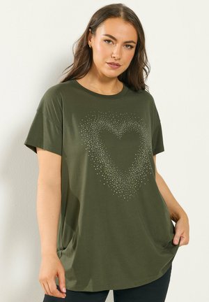 Frau trägt ein olivgrünes, übergroßes T-Shirt mit gepunktetem Herzdesign, posiert mit einer Hand, die den Saum hält, vor hellem Hintergrund.