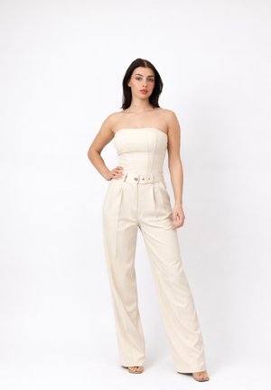 Vrouw met donker haar, gekleed in een crème kleurige strapless jumpsuit met ceintuur en open schoenen, staand tegen een effen witte achtergrond.