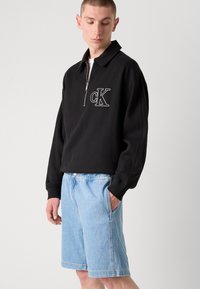Pullover nero a zip con collo a risvolto, caratterizzato da una texture a coste e un logo "CK" bianco sul petto, abbinato a un paio di shorts in denim blu chiaro.