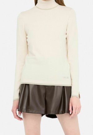 Femme portant un pull à col roulé crème et un short en cuir plissé marron foncé, se tenant contre un fond clair uni.