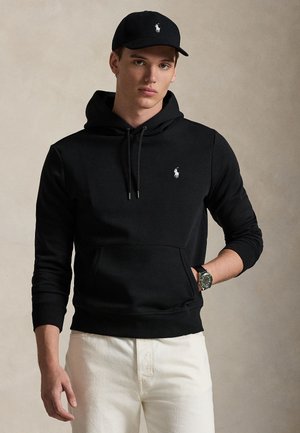 Jeune homme portant un sweat à capuche noir et une casquette noire assortie avec un logo blanc, pantalon blanc et montre-bracelet, debout sur un fond neutre.