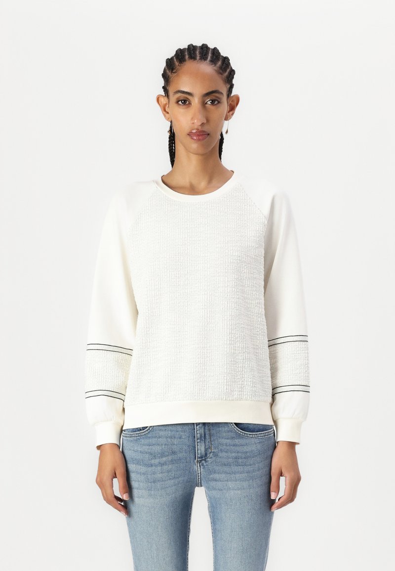 Valge sweatshirt tekstureeritud eesmine osa, mustad triibud varrukatel ja ribatud mansetid. Kombineeritud helesiniste teksadega.