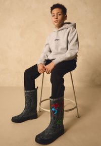 Svarta gummistövlar med Marvel-seriemönster, med Spider-Man och Hulk. Ett barn bär en grå huvtröja och svarta byxor, sitter på en metallpall.