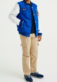 Modrá varsity bunda s bílými rukávy, se vzory a žebrovanými náplety. V kombinaci s béžovými cargo kalhotami a tmavě modrými teniskami.