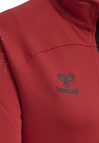 Rote Sportjacke aus strukturiertem Stoff, versehen mit einem hohen Kragen und einem schwarzen Reißverschluss. Das Hummel-Logo ist auf der Brust geprägt.