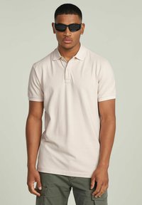 Polo shirt rosa chiaro in cotone, con colletto ribattuto e pattino a due bottoni. Indossato con occhiali da sole neri e pantaloni cargo scuri.