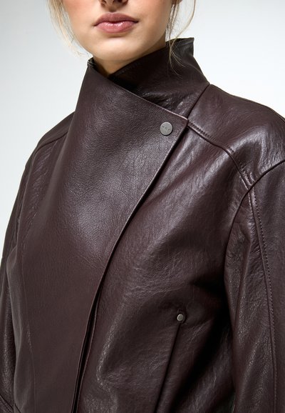 Nő sötétbarna bőrkabátot visel, amelynek magas gallérja és aszimmetrikus eleje van, fém patentokkal rögzítve.
