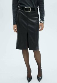 Jupe crayon en cuir noir avec une fente à l'avant, dotée d'une large ceinture avec une boucle en métal rectangulaire, portée avec des collants noirs transparents et des talons.