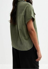 Kurzärmlige, olivgrüne Bluse aus einem weichen, strukturierten Stoff. Verfügt über einen lockeren Schnitt und einen abgerundeten Saum, ein Rückenpasse-Design und aufgerollte Ärmel.