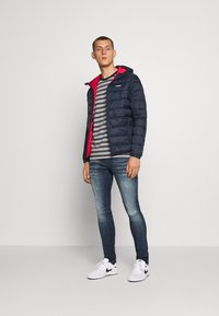 Marine blauwe pufferjack met een rode binnenkant, gestreept shirt eronder, blauwe vervaagde spijkerbroek en witte sneakers met zwarte accenten.
