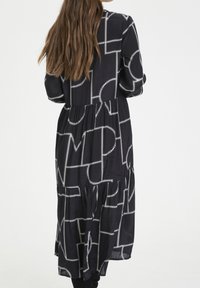 Mustapuku, jossa on valkoinen geometrinen kuvio, pitkät hihat, kerroksellinen muotoilu ja pehmeä, kevyt kangas. Näkyvä pituus nilkkoihin saakka.