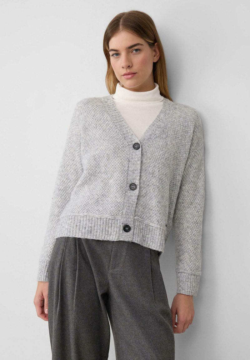 Grauer, strukturierter Cardigan mit V-Ausschnitt, vier schwarzen Knöpfen und geripptem Saum. Kombiniert mit hochtaillierten grauen Hosen. Glattes, weiches Material.