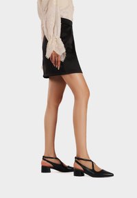 Scarpe in pelle nera con tacco a blocco e punta affusolata, con cinturini alla caviglia e un design elegante, indossate con una mini gonna nera e una blusa chiara.